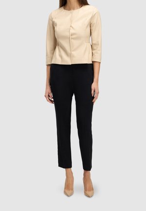 Femme portant une veste beige ajustée à manches trois-quarts, un pantalon noir slim et des escarpins beiges à bout pointu sur un fond uni.