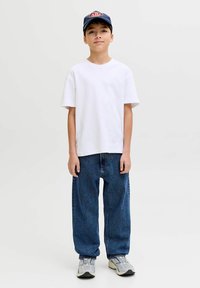 Fehér rövid ujjú póló laza sötétkék farmerrel és szürke tornacipővel, kiegészítve egy tengerészkék sapkával, amelyen piros logó található.