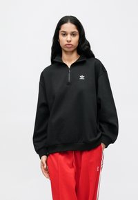 Juodas džemperis su užtrauktuku ir aukštu apykakliu, su mažu baltu Adidas logotipu. Lygios tekstūros ir laisvo kirpimo, derinamas su raudonomis kelnėmis.