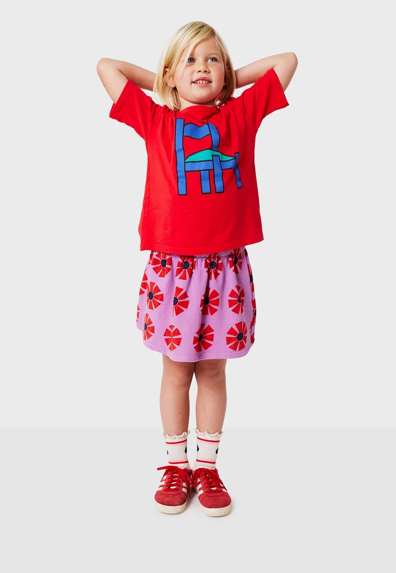 Rotes Baumwoll-T-Shirt mit einem blauen und grünen grafischen Design, kombiniert mit einem pinken Rock mit roten Blumen. Rote Sneakers mit weißen Socken.