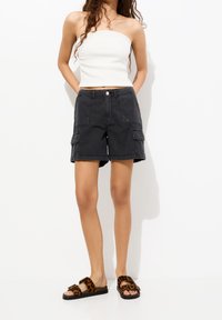 Pantalones cortos cargo negros con múltiples bolsillos, combinados con una blusa sin mangas blanca de canalé y sandalias marrones con estampado de leopardo. Textura suave.