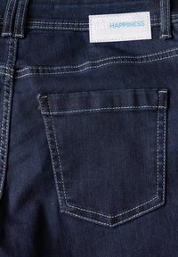 Vue arrière de jeans en denim foncé avec une seule poche arrière ornée de surpiqûres blanches et d'une étiquette portant la mention "PAS D'INQUIÉTUDES, JUSTE DU BONHEUR."