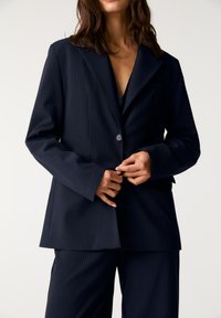 Marineblå blazer med smal stribe, enkelt knaplukning, brede revers og lige skåret silhuet. Stof er glat med en struktureret pasform.