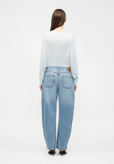 Kvinne med langt brunt hår iført en lyseblå langermet skjorte, høytlivede, løse blå jeans og svarte høye hæler, stående med ryggen til.