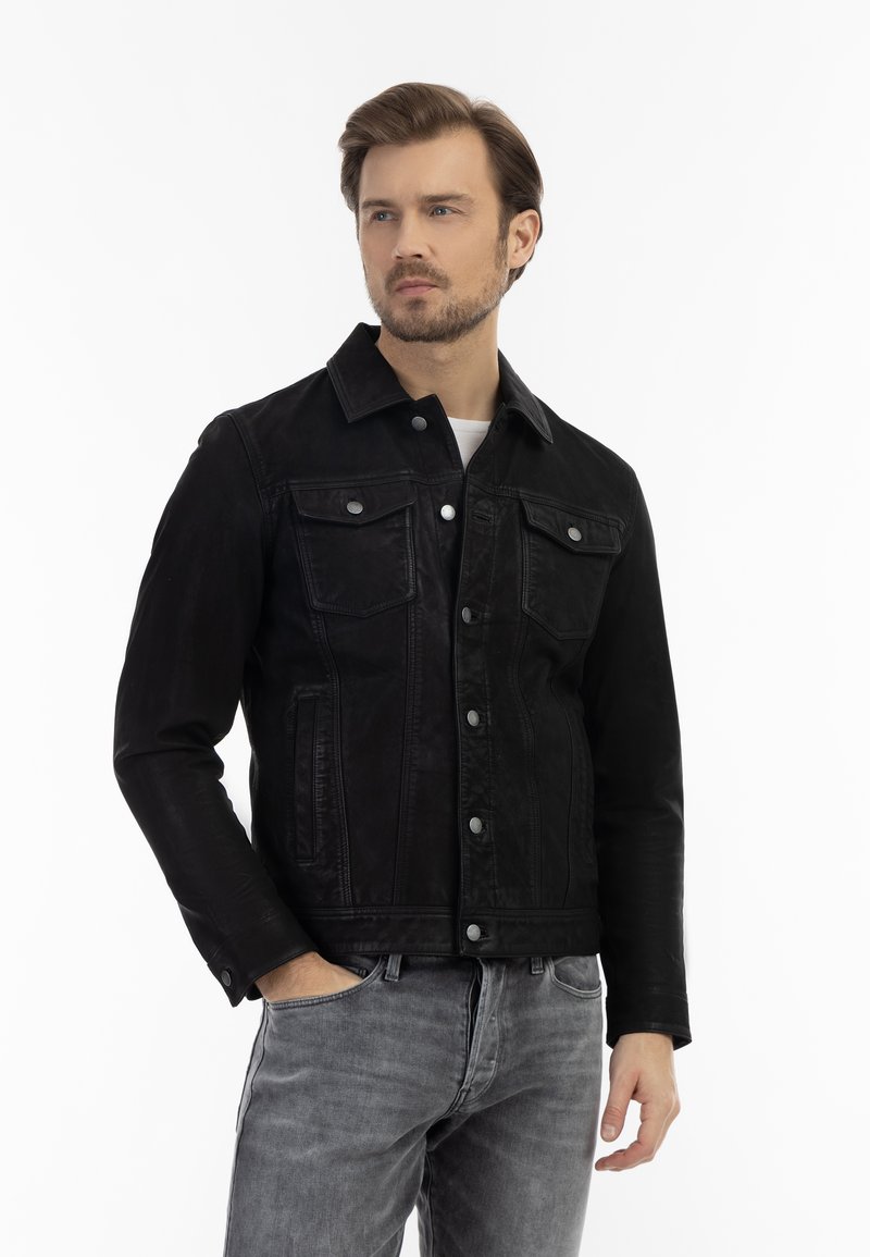 Jachetă din denim negru cu un design clasic, având buzunare frontale, nasturi metalici și un finisaj ușor texturat. Poartă o cămașă albă dedesubt.