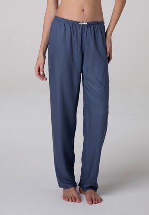 Donna che indossa pantaloni blu navy larghi con coulisse bianca, in piedi a piedi nudi su uno sfondo chiaro e semplice.