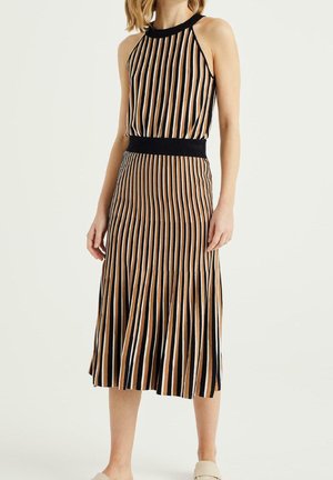 Femme portant une robe sans manches, longueur genou, à rayures verticales noires, beige et blanches, avec une ceinture noire, debout devant un fond uni.