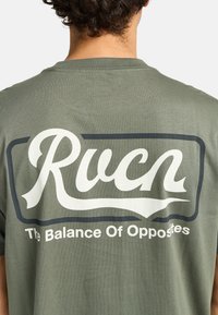 Olivegrünes Baumwoll-T-Shirt mit einem großen weißen "Rucn"-Logo und dem Text "The Balance Of Opposites" in einer kontrastierenden dunklen Umrandung.