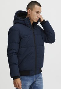 Marineblauwe pufferjas met capuchon, voorzien van een ritsluiting, een doorgestikt patroon, geribbelde manchetten en zijzakken. Draagt over een zwarte shirt.