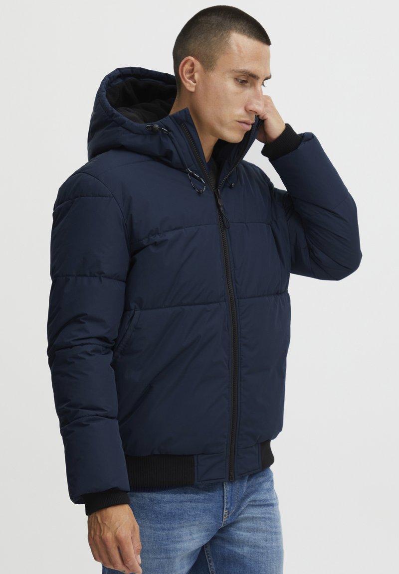 Marineblauwe pufferjas met capuchon, voorzien van een ritsluiting, een doorgestikt patroon, geribbelde manchetten en zijzakken. Draagt over een zwarte shirt.