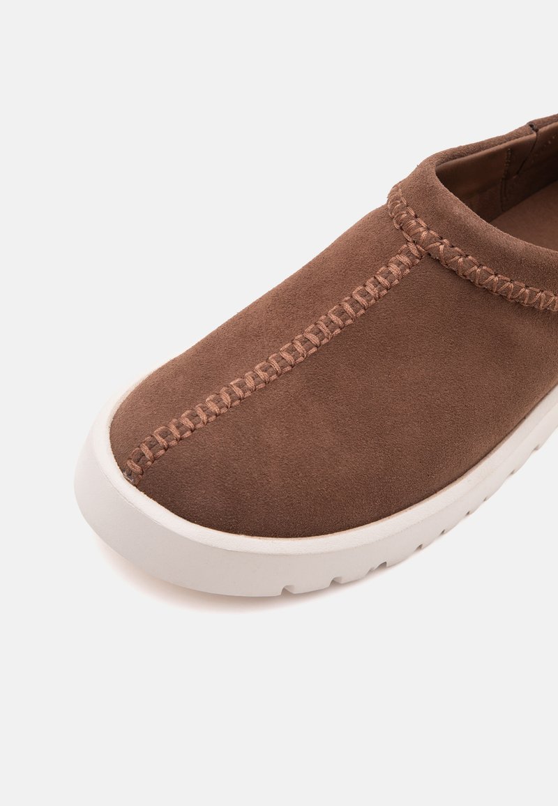 Zapato marrón de ante tipo slip-on con costuras decorativas a lo largo de la parte superior y suela de goma blanca sobre un fondo liso.