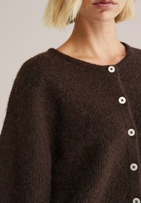 Barna kötött kardigán lekerekített nyakkal, nagy fehér gombokkal és texturált kivitelben. Puha, kényelmes anyag.