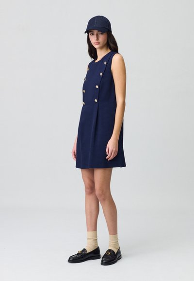 Vestido azul marino en corte A con botones dorados, diseño sin mangas y acentos plisados. Combinado con una gorra oscura, calcetas beige y mocasines negros.