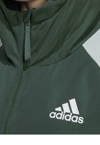 Tummanvihreä vetoketjullinen takki, jossa on korkea kaulus, vetoketjulla suljettava etuosa ja valkoinen Adidas-logo. Sileä, kevyt kangas, jossa on hienovarainen kiilto.