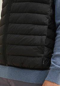 Schwarze Steppweste mit horizontalen Nähten, Reißverschluss und blauem geripptem Saum. Strukturiertes Material für Isolation und Wärme.