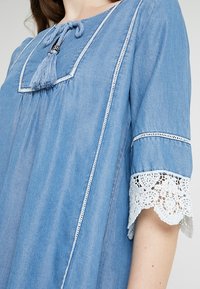 Světle modrá denimová tunika se čtvercovým výstřihem, ozdobným třásněmi, bílou krajkovou lemovkou na rukávech a podrobným prošíváním podél zapínání.