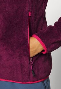 Chaqueta de forro polar en púrpura profundo con detalles en rosa. Cuenta con un cierre lateral y un bolsillo frontal con costuras visibles y tejido texturizado.