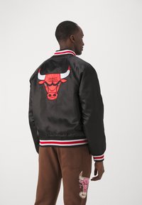 Svart bomberjacka med röd och vit randig ribbad krage. Har en stor röd tjurgraphic på baksidan. Bärs med bruna byxor.