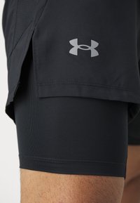 Černé sportovní šortky s perforovaným vzorem a elastickým střihem. Na boku se nachází stříbrné logo Under Armour. Hladká textura.