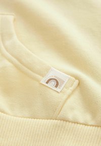 Tissu de sweatshirt jaune avec une texture lisse. Il est doté d'une poche kangourou et d'une étiquette avec un logo arc-en-ciel marron.