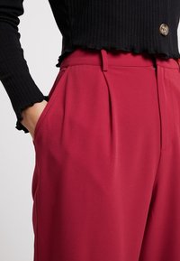 Rode broek van soepele stof met een hoge taille, voorzien van plooien en zijzakken, gecombineerd met een zwart geribbeld shirt met lange mouwen.