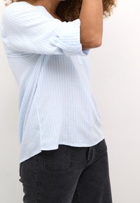 Chemise à rayures bleu clair avec manches retroussées, tissu doux, coupe décontractée, ourlet incurvé, associée à un jean foncé. Aucun motif ou embellissement visible.