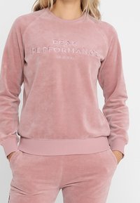 Sudadera de terciopelo rosa con puños y dobladillo acanalados. Presenta texto en relieve "PEAK PERFORMANCE ORIGINAL" en la parte frontal. Ajuste casual y relajado.