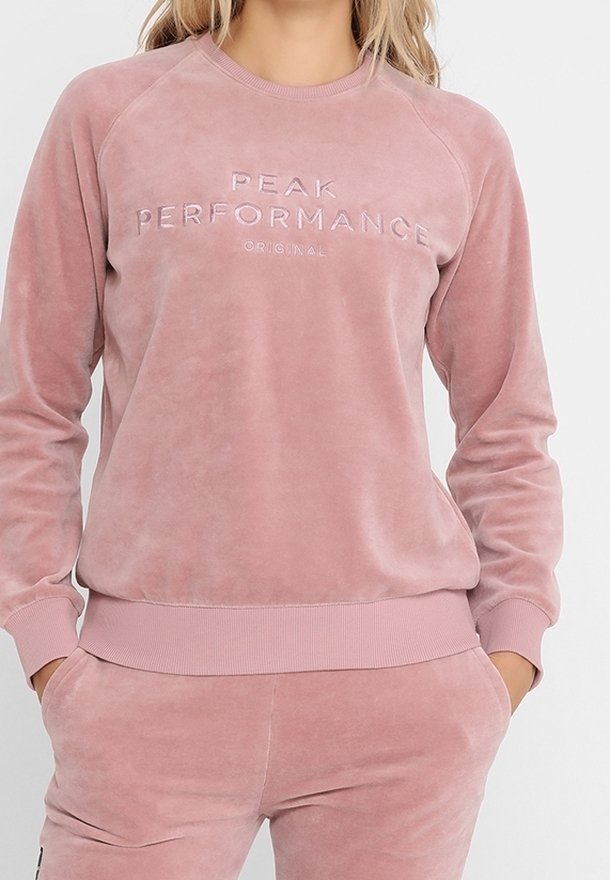 Sudadera de terciopelo rosa con puños y dobladillo acanalados. Presenta texto en relieve "PEAK PERFORMANCE ORIGINAL" en la parte frontal. Ajuste casual y relajado.