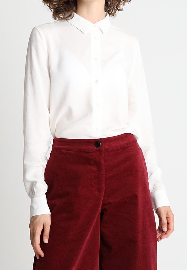 Camicia bianca con colletto, maniche lunghe e texture liscia, abbinata a pantaloni in velluto a coste color bordeaux con un motivo verticale.