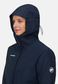 Donna che indossa una giacca Mammut blu navy con zip frontale e cordino per cappuccio regolabile, rivolta a sinistra su uno sfondo chiaro e semplice.