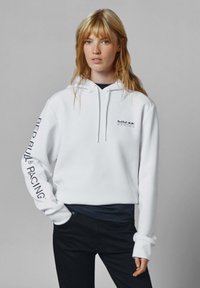 Blondínka s voľnými vlasmi, oblečená v bielom mikinovom svetri Red Bull Racing a čiernych nohaviciach, na jednotnom šedom pozadí.
