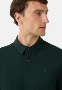 Dunkelgrünes Polohemd mit klassischem Kragen, dreiknöpfiger Blende und einem dezenten "B"-Logo auf der Brust. Strukturiertes Gewebe mit glatter Oberfläche.