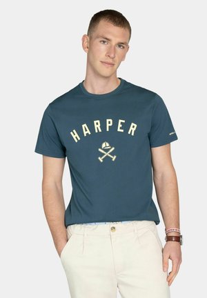 Joven con las manos en los bolsillos lleva una camiseta teal con la inscripción "HARPER" y un gráfico de bate y casco de béisbol cruzados, combinada con pantalones beige claros.