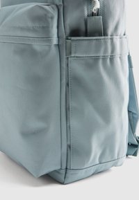 Ανοιχτό μπλε σακίδιο που διαθέτει υφή υφάσματος, πολλαπλές εξωτερικές τσέπες και πλαϊνή τσέπη με φερμουάρ. Ανθεκτικοί ιμάντες με λεπτομέρειες ενίσχυσης.