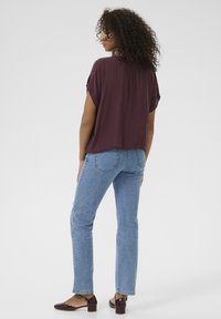Burgunderfarbenes, drapierenes Oberteil, kombiniert mit hellblauen, geraden Jeans. Lederschuhe mit niedrigem Blockabsatz und Knöchelriemen sind von hinten sichtbar.