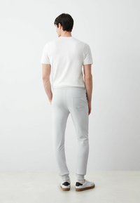 Gaiši pelēki sporta bikses ar elastīgiem materiāliem apakšgalos un aizmugurējo kabatu, saskaņotas ar baltu pieguļošu kreklu ar apaļu kakla izgriezumu un baltām kedām ar melniem akcentiem.