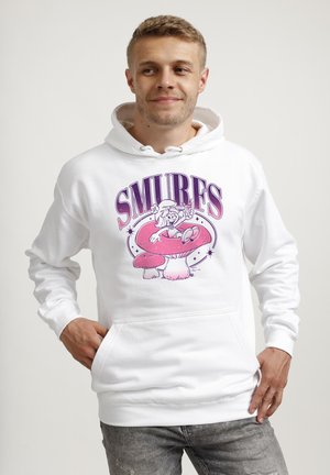 Młody mężczyzna w białej bluzie z kapturem z różową Smerfetką siedzącą na czerwonym grzybie i napisem „SMURFS” nad nim, lekko się uśmiecha.
