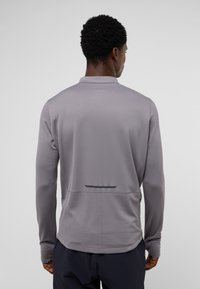 Lange mouwen, lichtgrijs sportshirt van ademend materiaal met een gestructureerde stof, kraag en een reflecterende strook aan de achterzijde. Heeft een ontspannen pasvorm en een detail aan de achternaad.