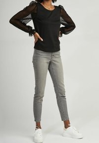 Top nero con maniche in pizzo trasparente caratterizzate da polsini a volant, abbinato a jeans skinny grigio chiaro e sneaker bianche. Design semplice e moderno.