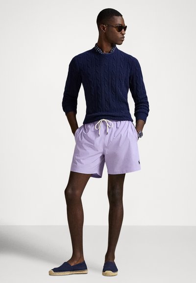 Temno moder pulover z vzorcem iz kaveljnih pletenin, bela srajca z ovratnikom, svetlo sivkaste kratke hlače in mornarsko modre espadrile; sončna očala dopolnjujejo izgled.