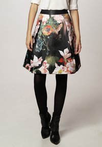 Jupe noire évasée avec un imprimé floral vif, présentant une taille ajustée et un design plissé. Portée avec des collants noirs et des bottines.