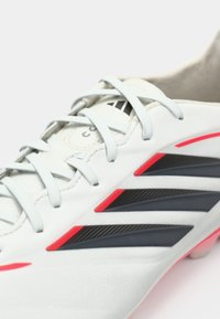 Λευκό ποδοσφαιρικό παπούτσι με γκρι λεπτομέρειες, διαθέτον τρεις μαύρες ρίγες και κόκκινες λεπτομέρειες. Ομαλή υφή με επίπεδα, λεπτά κορδόνια.