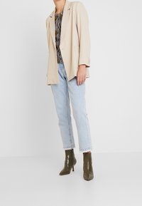 Beiger Blazer mit entspannter Passform und einem Knopf, kombiniert mit hellblauen geraden Jeans und dunkelgrünen, spitzen Knöchelschuhen.