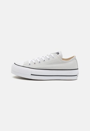 Witte lage canvas sneaker met witte veters, dikke witte rubberen zool en zwarte streepdetails op een effen achtergrond.