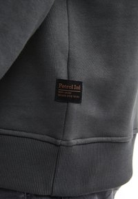 Felpa grigia scura con orlo e polsini a coste. Etichetta marrone con il testo "Petrol Ind" cucita sul lato. Tessuto di texture liscia.