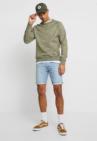 Felpa verde oliva con polsini a coste, abbinata a pantaloni shorts di jeans azzurri. Sneakers marroni con accenti bianchi e un cappellino verde indossati.