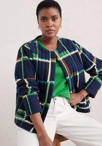 Pikowana granatowa kurtka z kolorowym wzorem w kratkę; zielony top pod spodem, białe spodnie. Ma zaokrąglony dekolt i mankiety z podwinięciem.