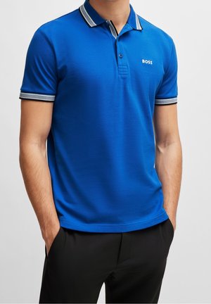 Mand iført en blå polo-shirt med hvide og grå kanter på krave og ærmer, hænder i lommerne og sorte bukser.