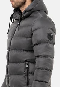 Graue Puffjacke mit Kapuze, mit einem Reißverschluss vorne, elastischen Kordeln und einem Patch am Ärmel. Sie hat seitliche Reißverschlusstaschen und ein gestepptes Design.