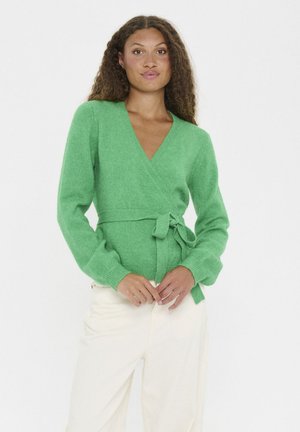 Vrouw met lang, krullend haar die een groene wikkeltrui draagt die op de taille is geknoopt, en off-white broek, staand tegen een witte achtergrond.